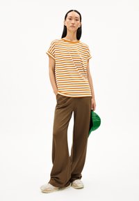 Striped t-shirt in orange and white, paired with brown wide-leg pants. The model holds a green cap and wears beige sneakers.