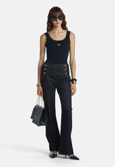 Kvinde iført sort ærmeløs top og sort bukser med høj talje og gyldne knapper, der holder en sort håndtaske med guldkæde, iført sorte hæle og solbriller.