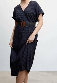 Laivastonsininen kietaisumekko, jossa on valkoisia pystysuoria raitoja. Lyhyet hihat ja syvä V-pääntie. Musta teksturoitu vyö, jossa on ruskea pyöreä solki vyötäröllä.