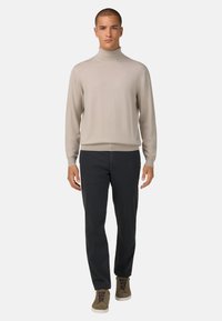 Svetlo bež turtleneck pulover, rebrasta tekstura, v kombinaciji s temno mornarskimi hlačami, rjavimi visoki čevlji z vezalkami, model stoji pokončno.