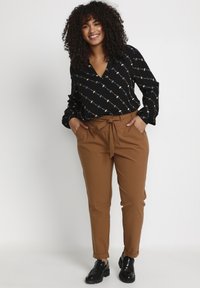 Black blouse with patterned figures, long sleeves, and a V-neck, paired with brown high-waisted trousers featuring a tied waistband and pockets.