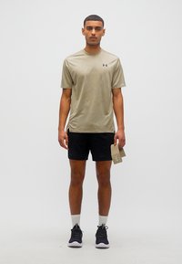 Άνδρας που στέκεται με το πρόσωπο προς τα εμπρός, φοράει μπεζ κοντομάνικο πουκάμισο, μαύρα σορτς, λευκές κάλτσες, μαύρα αθλητικά παπούτσια, κρατώντας μπεζ πορτοφόλι στο δεξί χέρι.