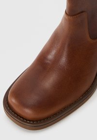 Bottine en cuir marron avec une texture lisse, à bout rond et couture contrastante le long de la semelle. Design minimaliste sans ornements.