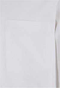 Tessuto di cotone bianco con una tasca rettangolare sul petto. La texture è liscia e morbida. La tasca presenta cuciture rinforzate intorno ai bordi.