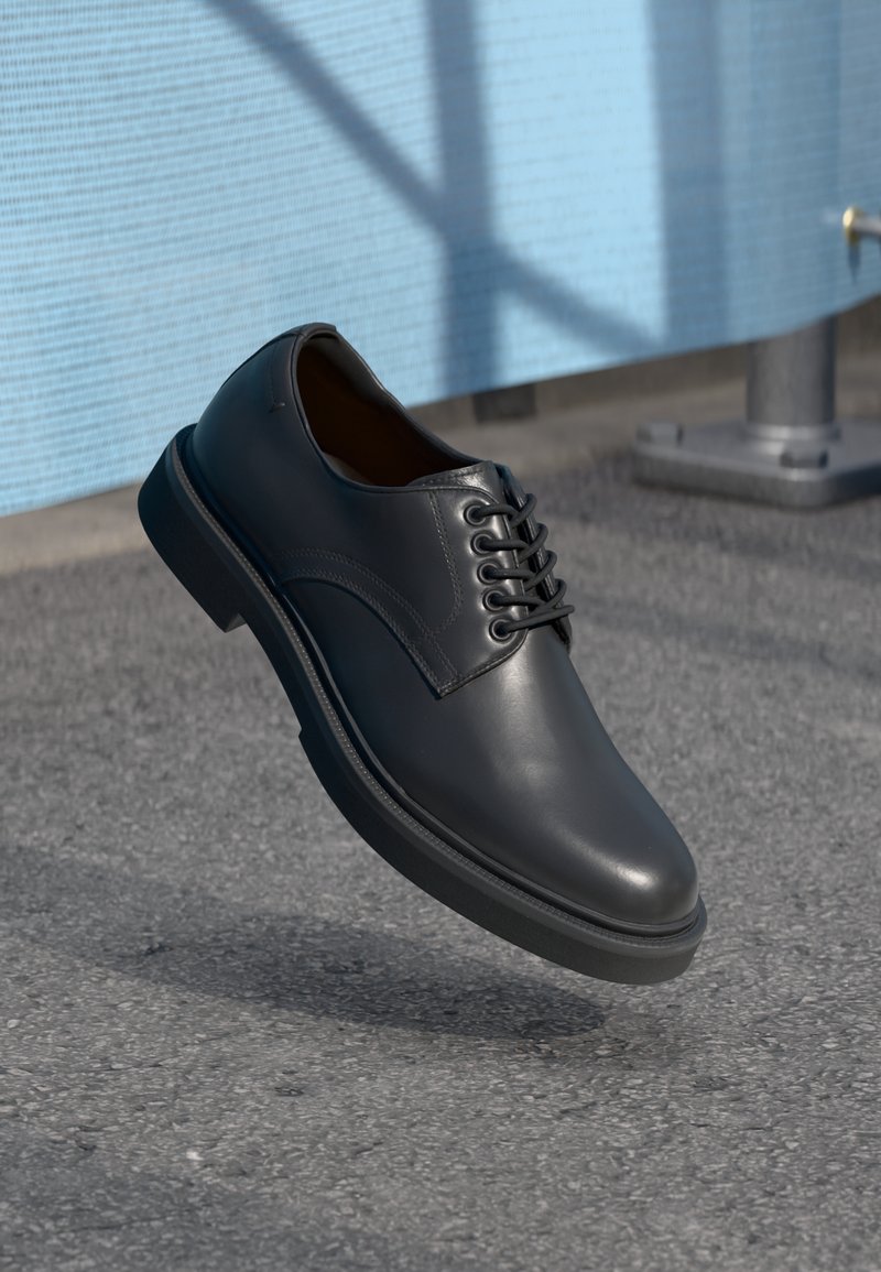 Chaussure habillée à lacets en cuir noir flottant au-dessus du sol en asphalte, avec un mur texturé bleu clair et un poteau métallique en arrière-plan.