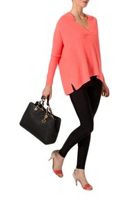Maglione a V in corallo, leggings neri e tacchi beige con cinturini rossi. Modella che tiene una borsa nera con accessori dorati.
