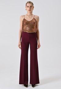 Donna che indossa un top senza maniche in bronzo con paillettes, pantaloni larghi bordeaux a vita alta, orecchini d'oro, collana e scarpe nere a punta.