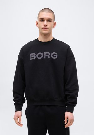 Young man with short hair wearing a black sweatshirt with "BORG" in large letters on the chest, standing against a plain background.