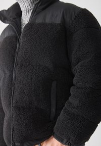Primo piano di una persona che indossa una giacca in pile nera con texture, dotata di cerniera, tasche a toppa e un maglione grigio a maglia sotto.