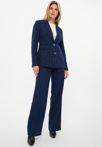 Traje de rayas azul marino que incluye un blazer ajustado con dos botones, bolsillos rectangulares y pantalones de pierna ancha en un material suave.