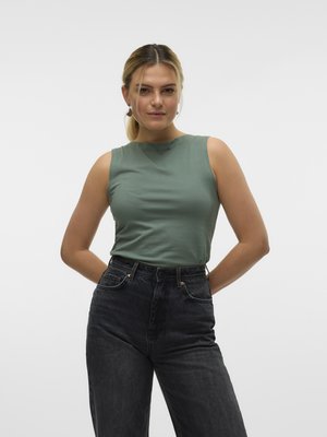 Femme aux cheveux blonds attachés en queue de cheval, portant un haut vert sans manches et un jean noir taille haute, debout, les mains derrière le dos.