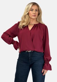Burgundowa bluzka z dekoltem w serek, długimi bufkami i elastycznymi mankietami, zestawiona z ciemnymi dżinsami z wysokim stanem. Złoty łańcuszek przy dekolcie.