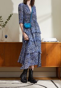 Maxi vestido azul con estampado y diseño en capas, combinado con un pequeño bolso bandolera turquesa y botas negras hasta el tobillo. Textura de tela suave.