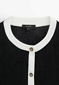 Cardigan en maille noire avec col blanc et patte de boutonnage ornée de boutons décoratifs. Le tissu est doux, léger et légèrement texturé.