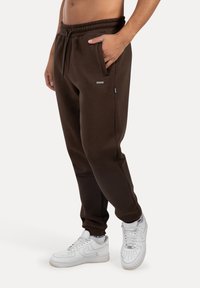 Mann trägt dunkelbraune Jogginghosen mit elastischem Bund und weißen Sneakers, steht mit einer Hand in der Tasche vor einem weißen Hintergrund.