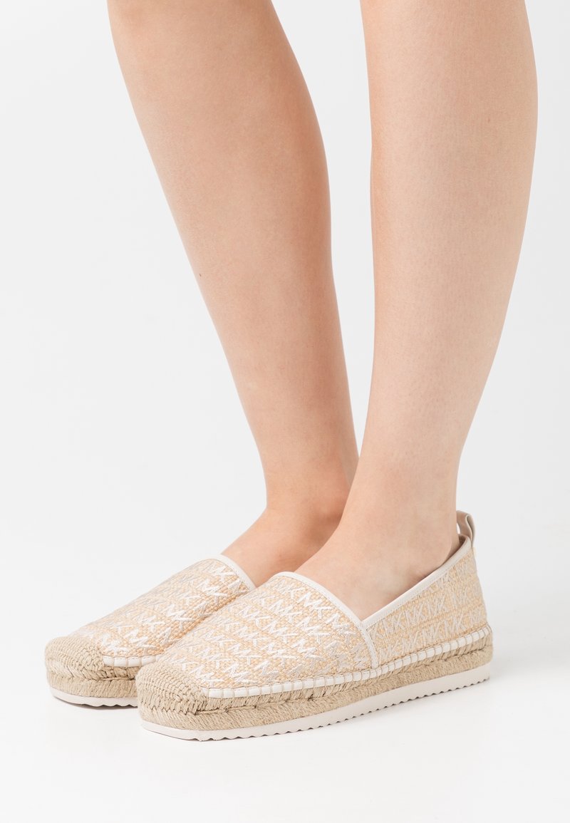 Pieds portant des espadrilles beiges tissées à enfiler avec des motifs en zigzag blanc et des semelles intermédiaires en corde de jute sur fond blanc.