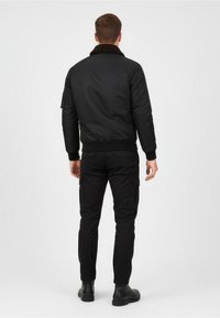 Giacca bomber nera con colletto in pelliccia marrone, texture trapuntata, polsini a coste e cintura a costine. Indossata con pantaloni cargo neri e stivali di pelle.