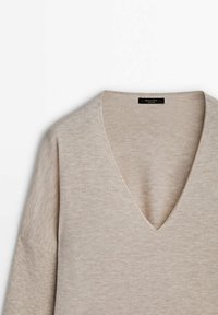 Pull en tricot beige avec un col en V, des manches larges et une texture douce. Il comporte une étiquette avec le nom de la marque à l'intérieur du col.