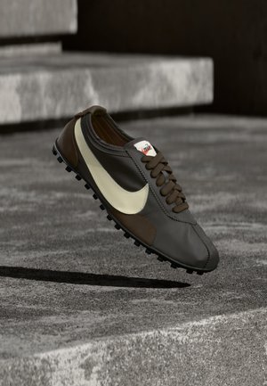Hnedý a béžový Nike teniska s ozubenou podrážkou vo vzduchu nad drsnými betónovými schodmi v neutrálnom vonkajšom prostredí.