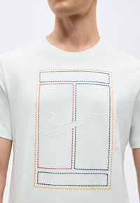 Άνδρας που φοράει ανοιχτόχρωμο πουκάμισο με ραμμένες πολύχρωμες ορθογώνιες περιγράμματα και λευκό σημειωμένο λογότυπο Nike swoosh στο κέντρο του στήθους.