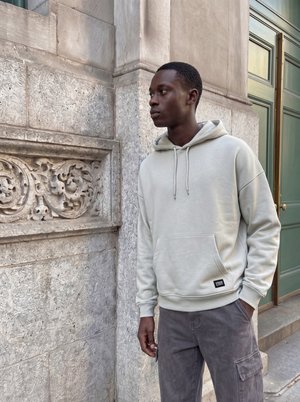Jeune homme en sweat à capuche beige clair et pantalon cargo gris debout à côté d’un mur de pierre orné et d’une porte verte, regardant sur le côté.