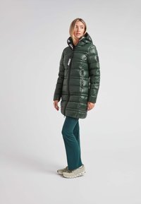 Chaqueta verde acolchada con un acabado brillante, cremallera negra y capucha con detalles de logo. Modelo vistiendo pantalones azul verdoso y zapatos de color claro.