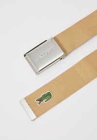 Cinturón de tela tejido en color beige con hebilla de metal plateado que presenta la grabación "LACOSTE"; detalle bordado del logo del cocodrilo en verde.