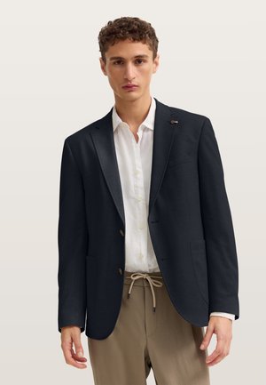 Junger Mann trägt einen schwarzen Blazer über einem weißen Hemd mit Knöpfen und beige Kordelzughosen vor einem schlichten hellen Hintergrund.