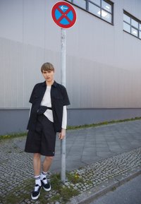 Chemise noire à manches courtes portée sur un haut en maille blanche, associée à un short noir. Baskets noires avec des accents blancs. Personnes près d'un panneau d'interdiction de stationner.