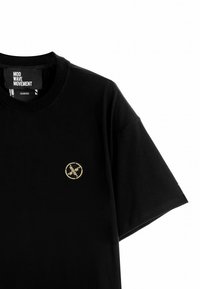 Camiseta de algodón negra con un logo bordado dorado redondo en el lado izquierdo del pecho. Mangas cortas y cuello redondo.