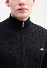 Fekete kötött, cipzáros pulóver, kábelmintával, magas bordás gallérral és logóval ellátott patch-csel. Elöl cipzárral és texturált anyaggal rendelkezik.