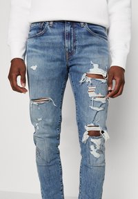 Jeans de mezclilla azul claro con detalles desgastados, que presentan grandes roturas, bordes deshilachados y un sutil desvanecimiento. Diseño estándar de cinco bolsillos.