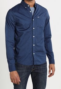 Man som bär en mörkblå skjorta med knappar över en grå t-shirt och mörkblå jeans, stående med armarna avslappnat vid sidorna.