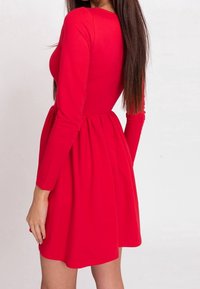 Czerwona sukienka z długim rękawem, wykonana z miękkiego materiału, z dopasowanym gorsetem i rozkloszowanym dołem. Widok z tyłu podkreśla szew i pas.