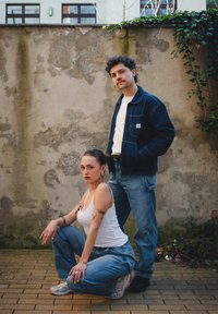 Due persone posano contro un muro testurizzato. Una indossa una giacca di jeans scura e jeans chiari, mentre l'altra ha un top bianco e jeans larghi.