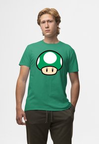 Zelené bavlněné tričko s velkým, stylizovaným zeleným grafickým motivem houbového klobouku se bílými tečkami a světlohnědým obličejem uprostřed.
