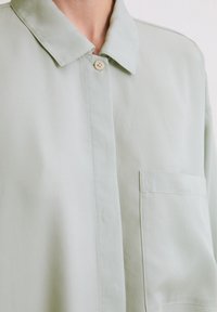 Light green button-up shirt with a collar, a single chest pocket, and smooth texture. Features minimal stitching and a relaxed fit.