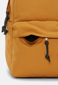 Sac à dos en tissu marron-orange avec une poche zippée. Doté de quincaillerie noire, d'une surface texturée et d'une fermeture à cordon.
