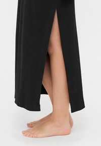 Vestido maxi negro de canalé con aberturas laterales y un corte holgado, con una textura suave y una longitud hasta los tobillos. Pies descalzos visibles.