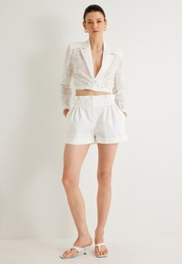 Vit kort blazer med blommönsterbroderi och djup V-ringning, tillsammans med vita plisserade shorts med sidfickor och uppvikt nederkant.