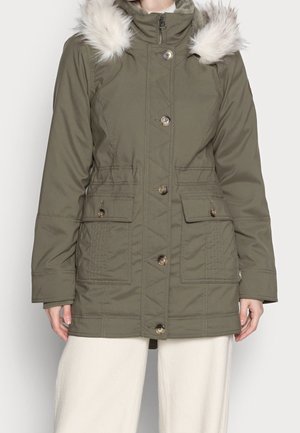 Mujer con parka de invierno verde oliva con capucha con borde de piel, cierre de botones y cremallera, y dos grandes bolsillos delanteros.