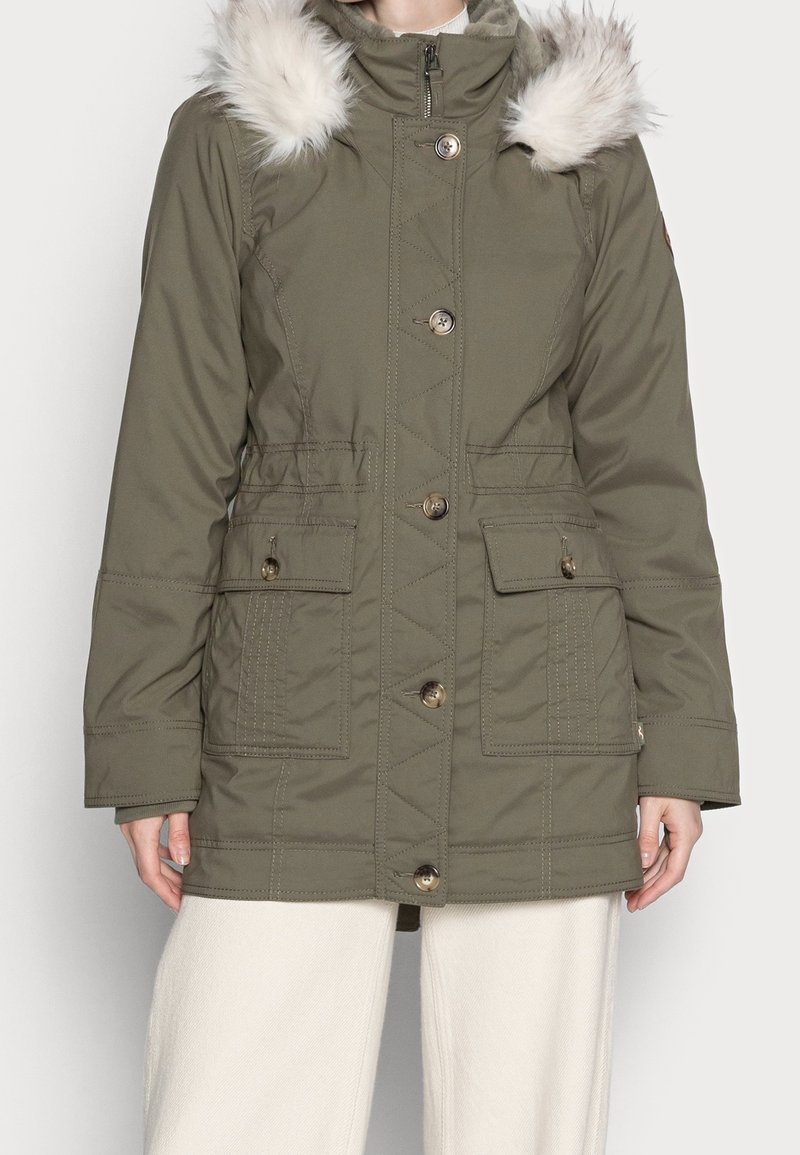 Mujer con parka de invierno verde oliva con capucha con borde de piel, cierre de botones y cremallera, y dos grandes bolsillos delanteros.