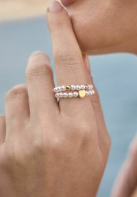 Tre staplade ringar: två gjorda av små vita pärlor och en guldring med en hjärtdekoration. Slät yta, delikat design, minimalistiska färger.