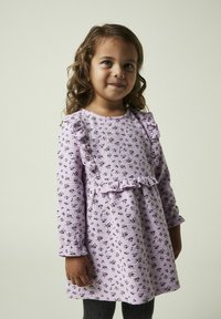 Lila långärmad klänning med volang på axlarna, texturerad tyg och blommönster. Kombinerad med grå leggings.