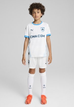 Maillot de football blanc avec des accents bleus et un logo, associé à un short blanc et des chaussettes montantes blanches. Des crampons orange vif complètent la tenue.
