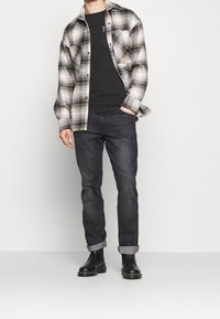 Uomo che indossa stivali neri, jeans scuri con risvolto, t-shirt nera e una camicia a quadri grigia e beige aperta, in piedi con una mano in tasca.