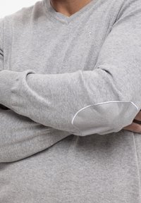 Personne portant un pull gris clair en maille avec des coudières cousues, bras croisés sur la poitrine.