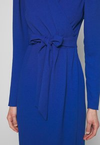 Robe bleue à manches longues avec une ceinture en tissu nouée en nœud à la taille, portée par une personne avec les mains visibles sur les côtés.