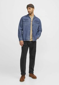 Denimjack met twee borstzakken, beige t-shirt eronder, zwarte jeans en bruine laarzen. Model draagt een bruine beanie.