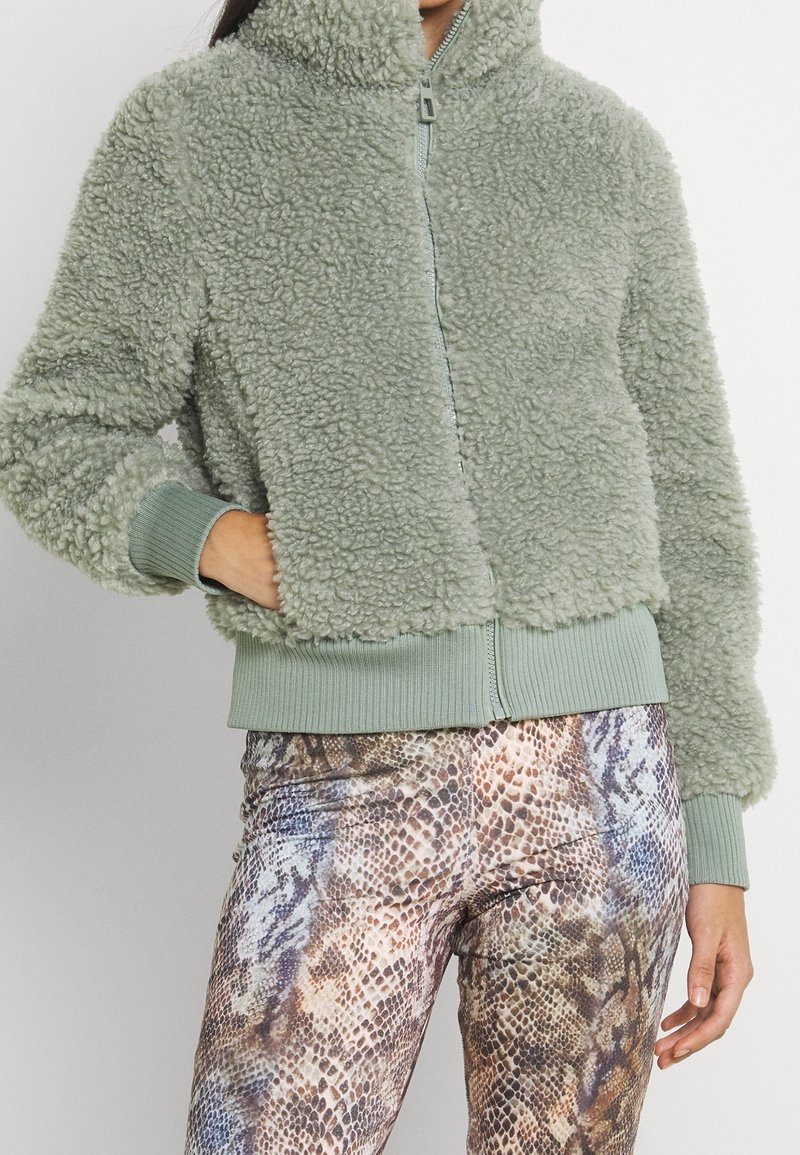 Femme portant une veste zippée légère vert clair, duveteuse, avec des poignets et un ourlet côtelés, associée à un pantalon taille haute imprimé serpent.